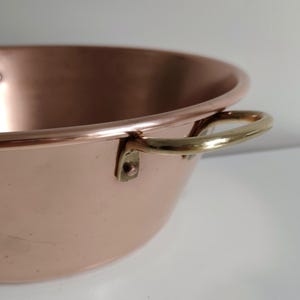 May include: A close-up shot of a copper bowl with brass handles. The bowl has a rounded shape and a polished finish, reflecting the light. The brass handles are attached to the sides of the bowl with rivets. The bowl is likely used for cooking or serving.