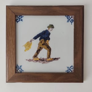 May include: A square ceramic tile framed in dark wood featuring a hand-painted illustration of a person in a green hat, blue shirt, and tan trousers walking while scattering yellow seeds. Blue floral corner accents decorate the white tile.