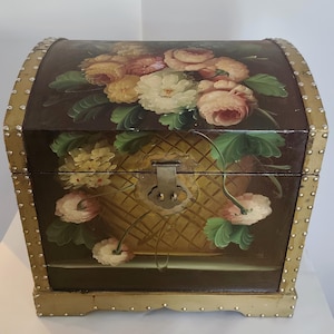 May include: A dark brown wooden chest with a domed lid, featuring hand-painted floral arrangements of pink, yellow, and white roses. The chest is accented with gold-toned metal trim along the edges, decorated with small, evenly spaced metal studs.