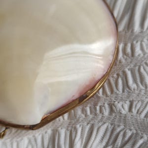 May include: A smooth, iridescent white and pale pink mother-of-pearl shell pendant or decorative piece encased in a thin, polished gold-toned metal frame. The surface of the shell displays a natural, glossy luster with subtle color variations.