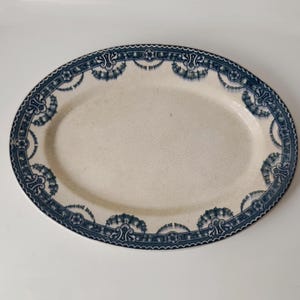 May include: An oval ceramic serving platter featuring a cream-coloured centre with a visible crackle glaze finish. The rim is decorated with a dark blue transferware pattern consisting of floral motifs, scrolling elements, and repeating geometric borders.