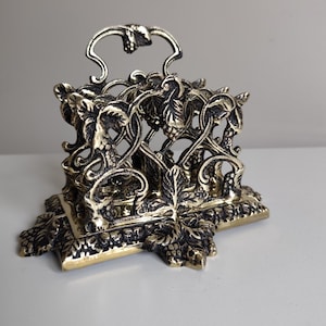 May include: An ornate brass napkin holder with a handle. The holder features a detailed design of grape vines and leaves. The base is square with decorative leaf accents. The overall colour is a mix of gold and dark bronze.