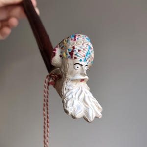 May include: A vintage tobacco pipe featuring a carved face with a long white beard and a colorful turban. The pipe has a dark brown stem and a red and white cord attached. The face has a stern expression.