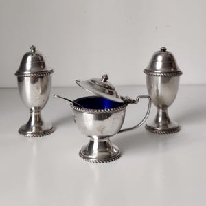 Puede incluir: Un juego de tres recipientes para condimentos de metal plateado que consta de dos saleros con tapas perforadas y un recipiente abierto con tapa abatible, asa y una cuchara pequeña, con interior azul profundo y borde trenzado.