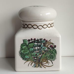 Può includere: Barattolo in ceramica bianca con coperchio e un disegno floreale di erbe verdi e fiori rosa. Il barattolo ha un disegno a corda marrone intorno alla parte superiore.