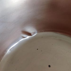 May include: Close-up of a shiny, metallic surface with a noticeable dent. The surface reflects light, creating bright highlights and shadows. The overall colour is a muted silver with a slight reddish hue in some areas. Small dark spots are scattered across the surface.