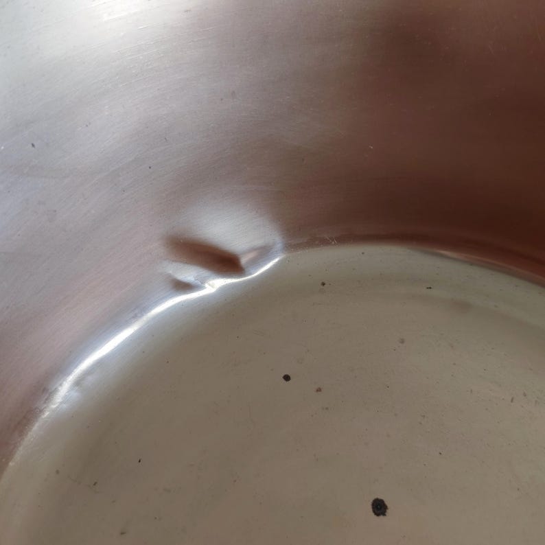 May include: Close-up of a shiny, metallic surface with a noticeable dent. The surface reflects light, creating bright highlights and shadows. The overall colour is a muted silver with a slight reddish hue in some areas. Small dark spots are scattered across the surface.