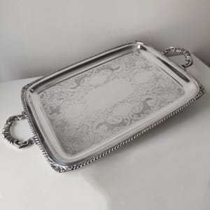 Puede incluir: Una bandeja de servicio rectangular de tono plateado con un patrón floral grabado ornamentado en el centro, un borde liso pulido, un borde estilo cuerda trenzada y dos asas curvas decorativas en los lados cortos de la bandeja.