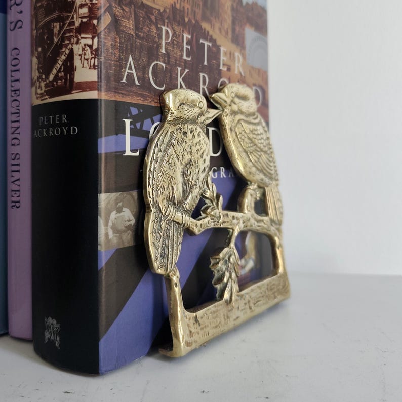 May include: A brass bookend featuring two birds perched on a branch with leaves. The bookend is positioned against a book titled London by Peter Ackroyd, with another book titled Collecting Silver visible on the left side of the frame.