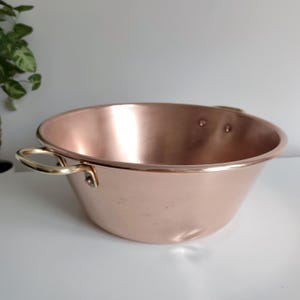 May include: A copper bowl with brass handles. The bowl has a rounded shape with a wide opening and a polished finish. The brass handles are attached to the sides of the bowl. The bowl is sitting on a white surface.
