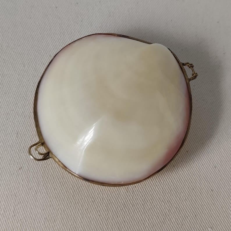 May include: A round, polished white seashell pendant with a smooth, iridescent surface and a subtle pink tint along the edge. The shell is encased in a thin, metallic gold-toned frame featuring two small loops on opposite sides for attachment.