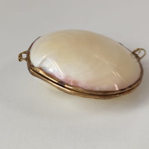 May include: An oval-shaped locket featuring a smooth, iridescent mother-of-pearl shell front encased in a thin, tarnished brass metal frame. The locket includes small metal loops on both sides for attachment, showcasing a natural, glossy finish.
