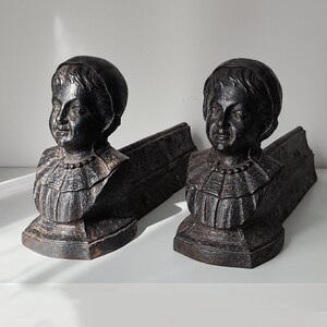 May include: A pair of dark, textured cast iron andirons featuring the sculpted busts of individuals wearing rounded caps and ruffled collars. Each piece has a sturdy, elongated base extending backward, designed for supporting logs in a fireplace.