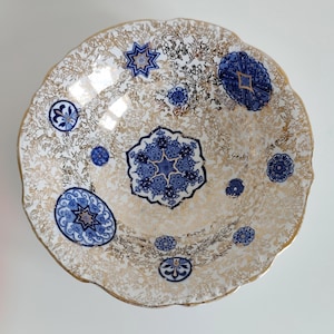 Puede incluir: Un cuenco decorativo con borde festoneado y un patrón floral dorado y blanco. El cuenco presenta diseños azules de estrellas y flores. El borde está adornado con oro. El cuenco es probablemente de cerámica o porcelana.