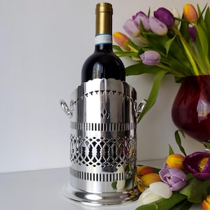 May include: A silver wine bottle holder with a decorative openwork design, holding a bottle of red wine. The holder has a scalloped top edge and small handles. A bouquet of colorful tulips is in the background.