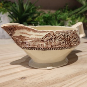 Pode incluir: Um molho de cerâmica marrom e branco com um design de cena de campo. O molho tem uma borda ondulada e uma alça.