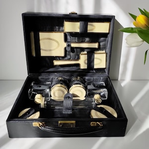 Puede incluir: Un neceser de cuero negro vintage, abierto para revelar una colección de accesorios de aseo de color marfil y dorado. El estuche incluye cepillos, polveras y frascos de perfume, todos cuidadosamente dispuestos. El estuche se cierra con un cierre dorado.