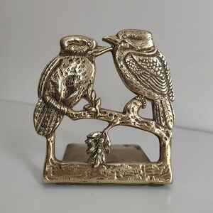 May include: A polished brass figurine featuring two kookaburras perched on a textured tree branch. The birds are facing each other with detailed feather engravings and defined beaks. The base is rectangular and supports the branch structure.