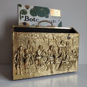 May include: A gold metal log holder with an embossed design of people gathered around a table. The log holder has a black felt lining and a brass handle. A book titled "Botanica" is sitting on top of the log holder.