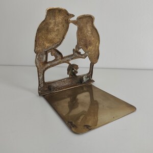 May include: A brass bookend featuring a cutout design of two birds perched on a branch. The vertical panel displays the bird silhouettes, while the horizontal base provides a flat, reflective surface for supporting books on a shelf.