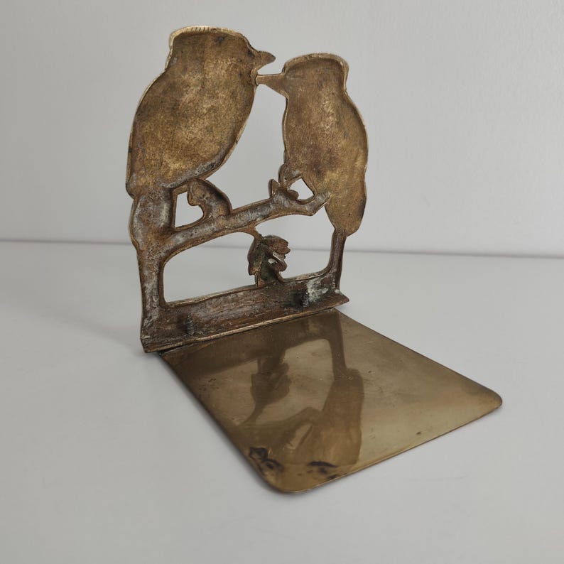 May include: A brass bookend featuring a cutout design of two birds perched on a branch. The vertical panel displays the bird silhouettes, while the horizontal base provides a flat, reflective surface for supporting books on a shelf.