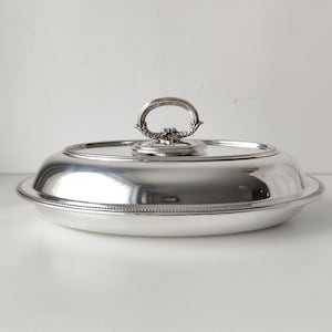 Puede incluir: Una fuente de servir ovalada en tono plateado pulido con tapa a juego. La tapa presenta un asa decorativa y ornamentada con detalles intrincados, y un borde con cuentas recorre el contorno tanto de la tapa como de la base de la fuente.