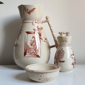 Può includere: Un set di tre pezzi in ceramica vintage di colore crema con motivi floreali e geometrici marroni. Il set comprende una grande brocca con manico, una brocca più piccola con manico e una piccola ciotola.