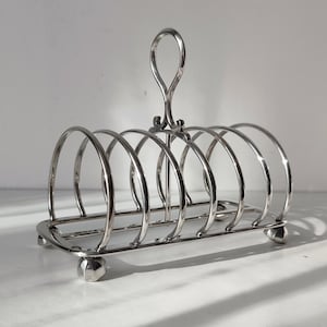 May include: A silver-toned metal toast rack featuring six arched slots for holding slices of bread. The rack has a central vertical handle with a loop at the top and is supported by four small, rounded feet at the base of the rectangular frame.