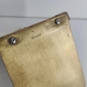 May include: A rectangular brass object with a brushed metallic finish. The surface features two silver-colored metal screws and stamped text that reads LOMBARD and MADE IN ENGLAND. The item has a flat, angled orientation with visible surface wear.