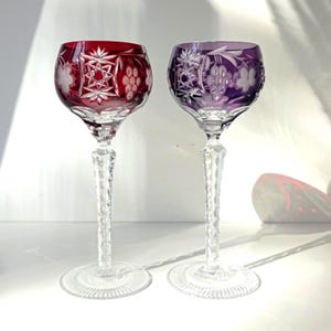 Könnte beinhalten: Zwei Weingläser aus Kristall mit klaren Stielen und Füßen. Ein Glas hat eine tiefrote Kuppa, das andere eine violette. Beide Kuppen sind mit aufwendigen Schliffmustern verziert, darunter sternförmige Motive und eingravierte Weintrauben.