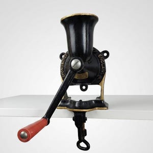 May include: A vintage cast iron hand-crank food grinder with a black body and gold-toned accents. The device features a red handle grip and is embossed with the text SPONG & CO LTD ENGLAND. It includes a top hopper and a bottom clamp mechanism.
