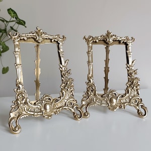 May include: A pair of antique-style picture frames in a gold finish. The frames feature elaborate scroll and floral designs, with a rectangular aperture for displaying images. The frames are made of metal and are designed to stand on a surface.