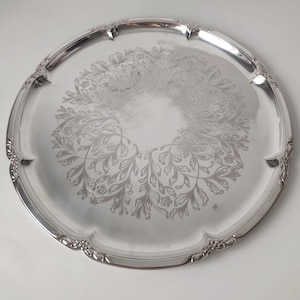 May include: A round silver-coloured metal serving tray featuring a polished finish and a decorative engraved floral pattern in the centre. The tray has a scalloped edge with embossed leaf and vine details along the raised rim of the platter.