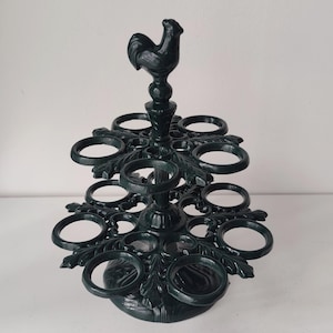 Op de afbeelding: Een donkergroene eierhouder van gegoten metaal met twee niveaus van cirkelvormige ringen voor het bewaren van eieren. De centrale verticale paal is bekroond met een decoratief haanfiguurtje. De basis en niveaus zijn versierd met bladpatronen.