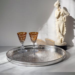 Könnte beinhalten: Ein rundes silberfarbenes Metalltablett mit einem komplizierten geprägten Blumenmuster und einem durchbrochenen Gitterrand, begleitet von zwei bernsteinfarbenen Kristallkelchen mit Diamantschliff und klaren Glasstielen auf einer Fläche.