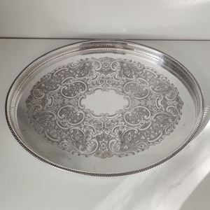 Könnte beinhalten: Ein ovales silberfarbenes Serviertablett mit einem komplizierten, geprägten Schnörkelmuster auf der Oberfläche. Das Tablett hat eine glatte, reflektierende Mitte und einen dekorativen, strukturierten Rand mit einem seilartigen Detail am gesamten Rand.