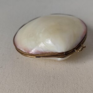 May include: A small, oval-shaped hinged box crafted from iridescent mother-of-pearl shell. The exterior features a smooth, creamy white surface with subtle pinkish hues along the edges, secured by a thin, metallic gold-toned brass frame and hinge.