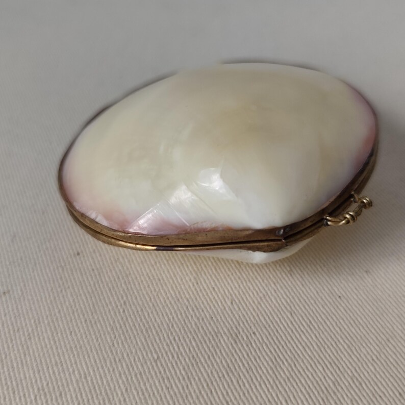 May include: A small, oval-shaped hinged box crafted from iridescent mother-of-pearl shell. The exterior features a smooth, creamy white surface with subtle pinkish hues along the edges, secured by a thin, metallic gold-toned brass frame and hinge.