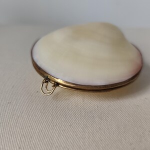 May include: A locket featuring a smooth, off-white seashell top set into a circular brass frame. The metal casing has a warm, aged patina and includes a small, double-looped brass bail attached to the side for hanging or attachment purposes.