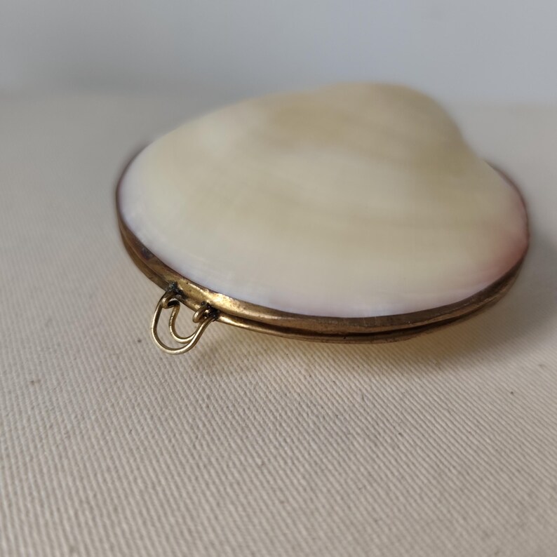 May include: A locket featuring a smooth, off-white seashell top set into a circular brass frame. The metal casing has a warm, aged patina and includes a small, double-looped brass bail attached to the side for hanging or attachment purposes.