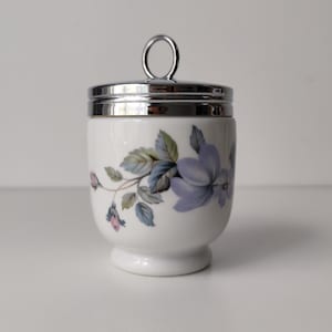 Może przedstawiać: Biały porcelanowy pojemnik do gotowania jajek z kwiatowym wzorem przedstawiającym niebieskie i fioletowe kwiaty oraz zielone liście. Pojemnik posiada błyszczącą, srebrną metalową pokrywkę z okrągłym uchwytem i zaokrągloną podstawę.