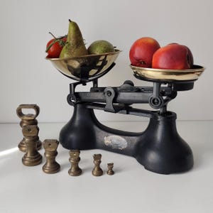 May include: A black cast iron balance scale with two brass pans. One pan holds a green pear and a red tomato, while the other holds two red nectarines. A set of six graduated brass weights sits in front of the scale, ranging from large to small.