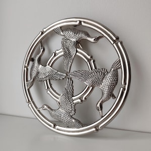May include: Circular silver-toned metal decorative piece featuring four stylized birds in flight. The birds are arranged around a central circle, with a bamboo-style border. The birds have detailed feather patterns and are in various poses.