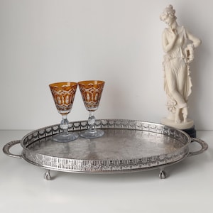 Peut inclure: Un plateau de service ovale en métal argenté avec un bord ajouré orné, deux poignées latérales et des pieds griffes. Deux verres en cristal ambré taillé avec des motifs géométriques complexes sont posés sur le plateau.