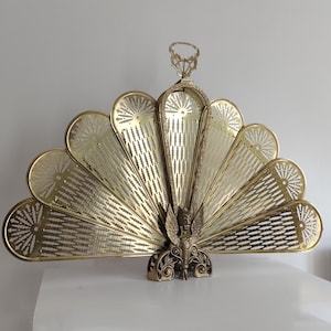 May include: A decorative brass fireplace screen in a fan shape. The screen features a detailed design with a central figure and intricate cut-out patterns. The brass has a warm, golden hue, adding a touch of elegance to any room.