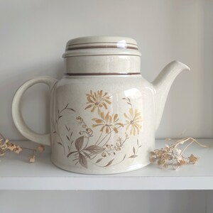 May include: A beige ceramic teapot with a brown lid and floral design. The teapot features a brown trim around the lid and a floral pattern with brown stems and yellow flowers.