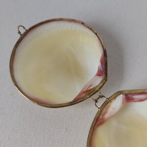 May include: An open circular locket or container made from two polished seashell halves joined by a thin metal hinge. The interior of the shells is smooth and creamy white with subtle pinkish-red edges, framed by a thin, tarnished gold-tone metal rim.
