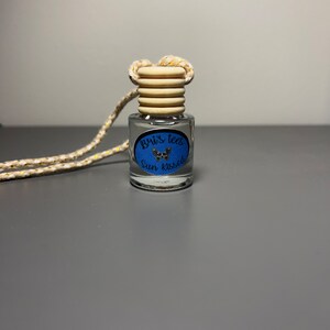 Op de afbeelding: Een klein glazen flesje met een houten dop en een blauw en gouden etiket met de tekst "Brix Tees Sun Kissed". Het flesje hangt aan een wit touwtje met gouden accenten.