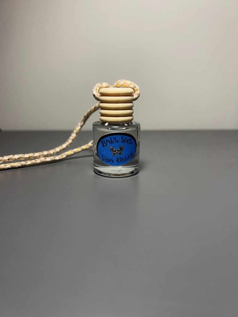 Op de afbeelding: Een klein glazen flesje met een houten dop en een blauw en gouden etiket met de tekst "Brix Tees Sun Kissed". Het flesje hangt aan een wit touwtje met gouden accenten.