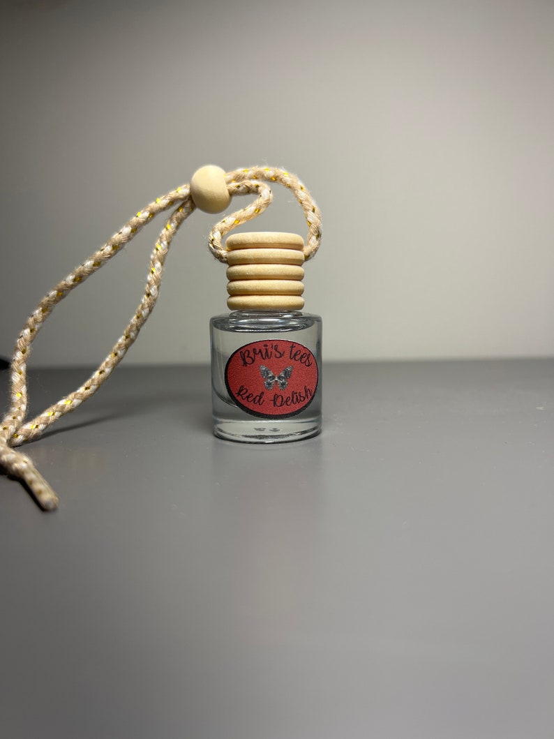 Op de afbeelding: Een doorzichtige glazen fles met een rood en wit etiket waarop "Bri's Tees Red + Delish" staat. De fles heeft een houten dop en een beige touwtje.